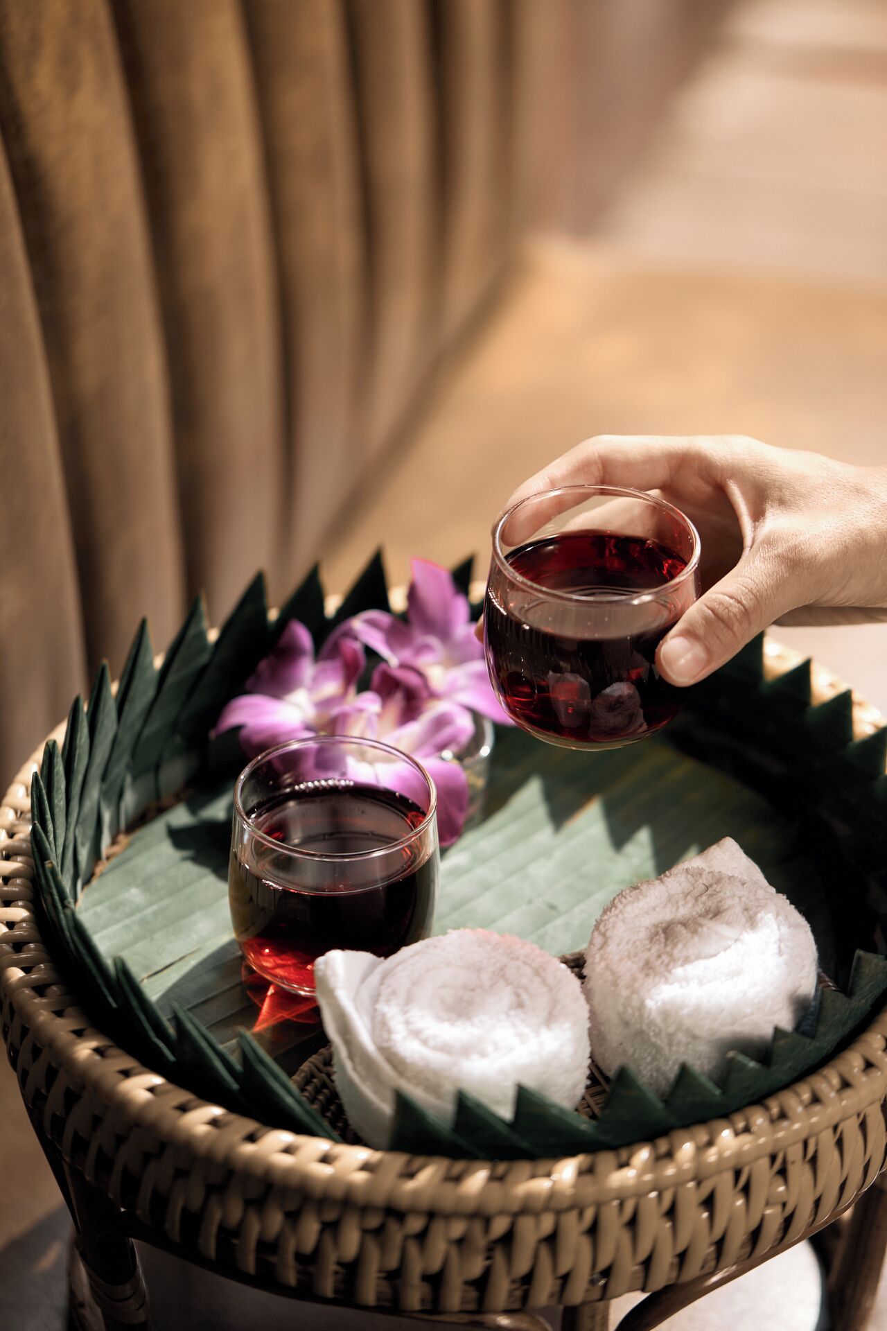 A hand holding a small glass of red liquid over a woven tray containing two white towels and two glasses of red liquid.