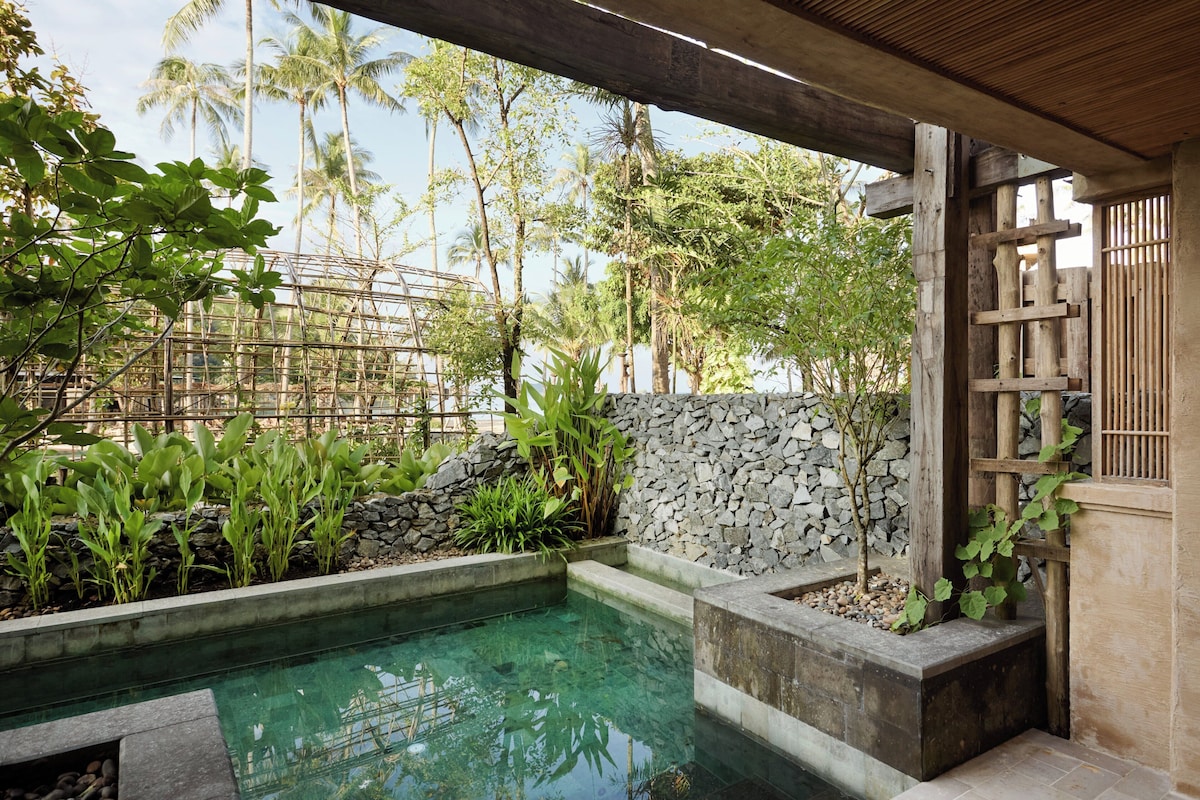 Ein klares Schwimmbecken mit Steinmauern und tropischen Pflanzen, umgeben von einer hölzernen Pergola und einem Bambuszaun.