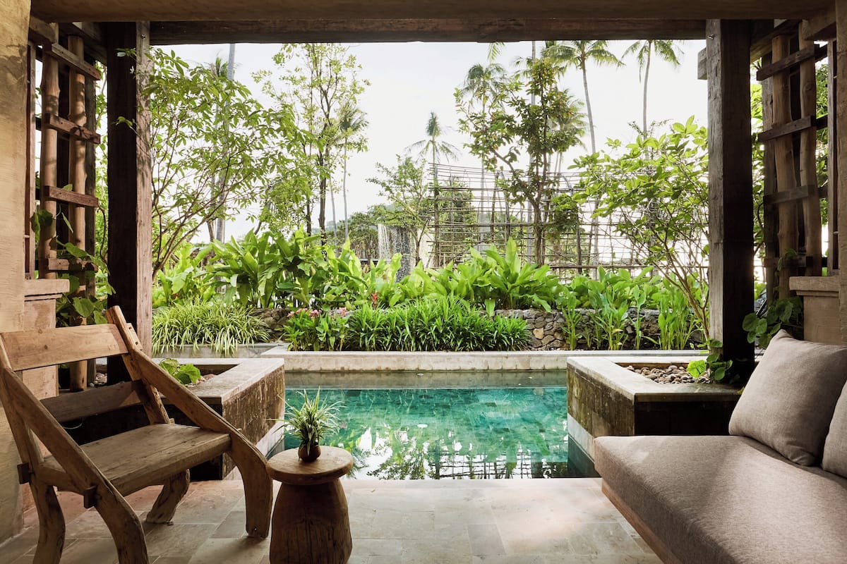 A pool area with a wooden bench and a cushioned seating area, surrounded by lush greenery and a view of palm trees.