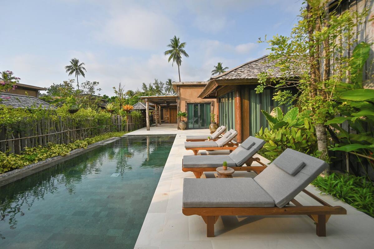 A pool area with lounge chairs and a clear pool, surrounded by greenery and palm trees.