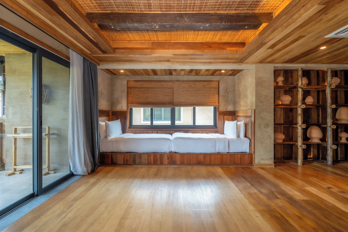 Ein Zimmer mit Holzboden, einem großen Fenster mit Jalousien, einer eingebauten Sitzbank mit Kissen und einem Regal mit Keramik.
