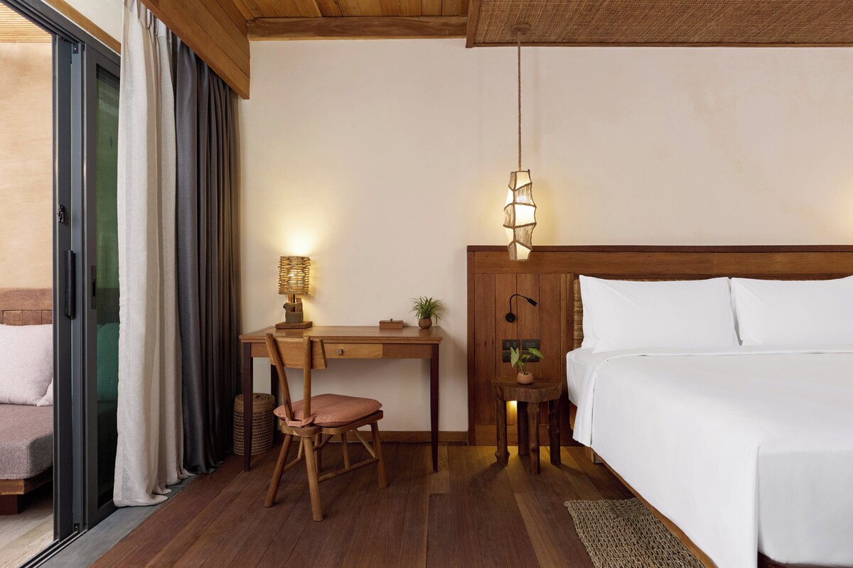 A hotel room with a bed, desk, chair, and a hanging light fixture.