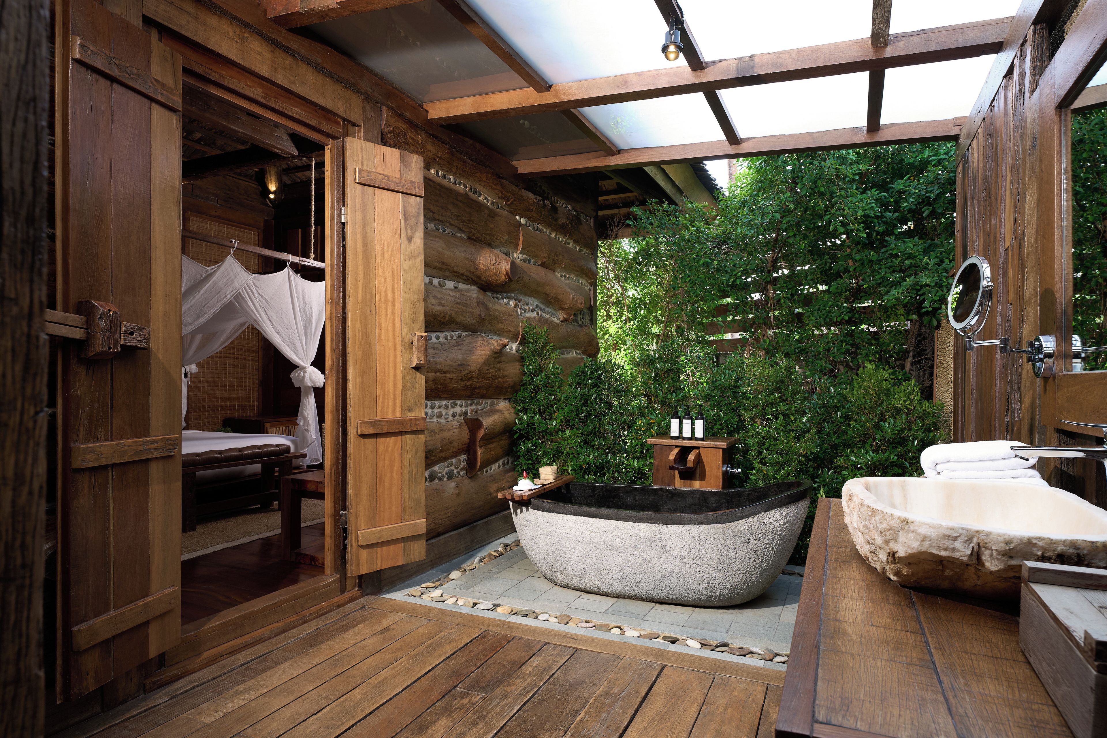 A log cabin interior with a wooden floor, a stone bathtub, a wooden bench, and a bed with a canopy.