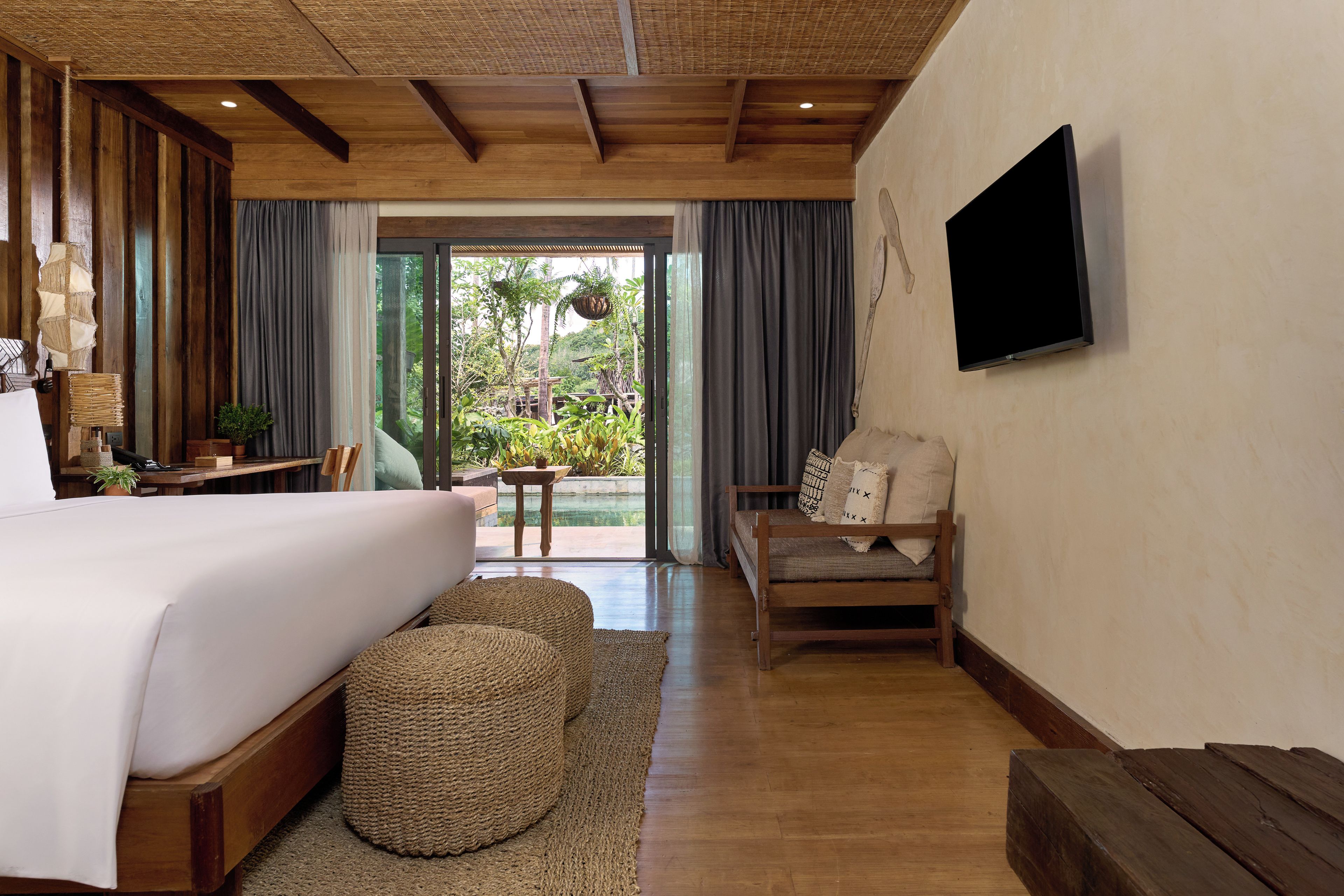 A bedroom with a large bed, a wooden floor, a television on the wall, and a view of a garden through the window.