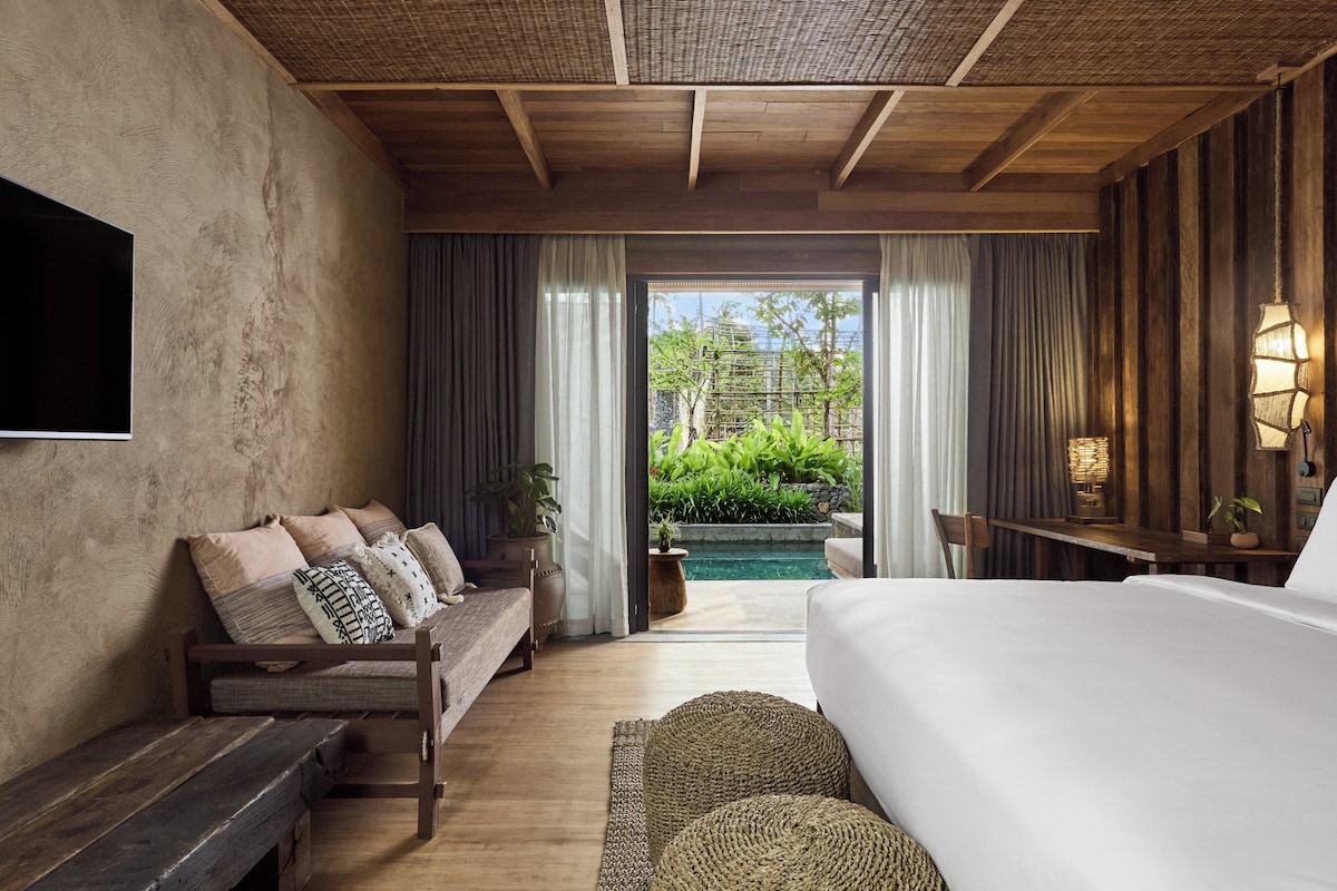 A bedroom with a large bed, a wooden bench, a sofa with cushions, a TV on the wall, and a view of a swimming pool and greenery outside.