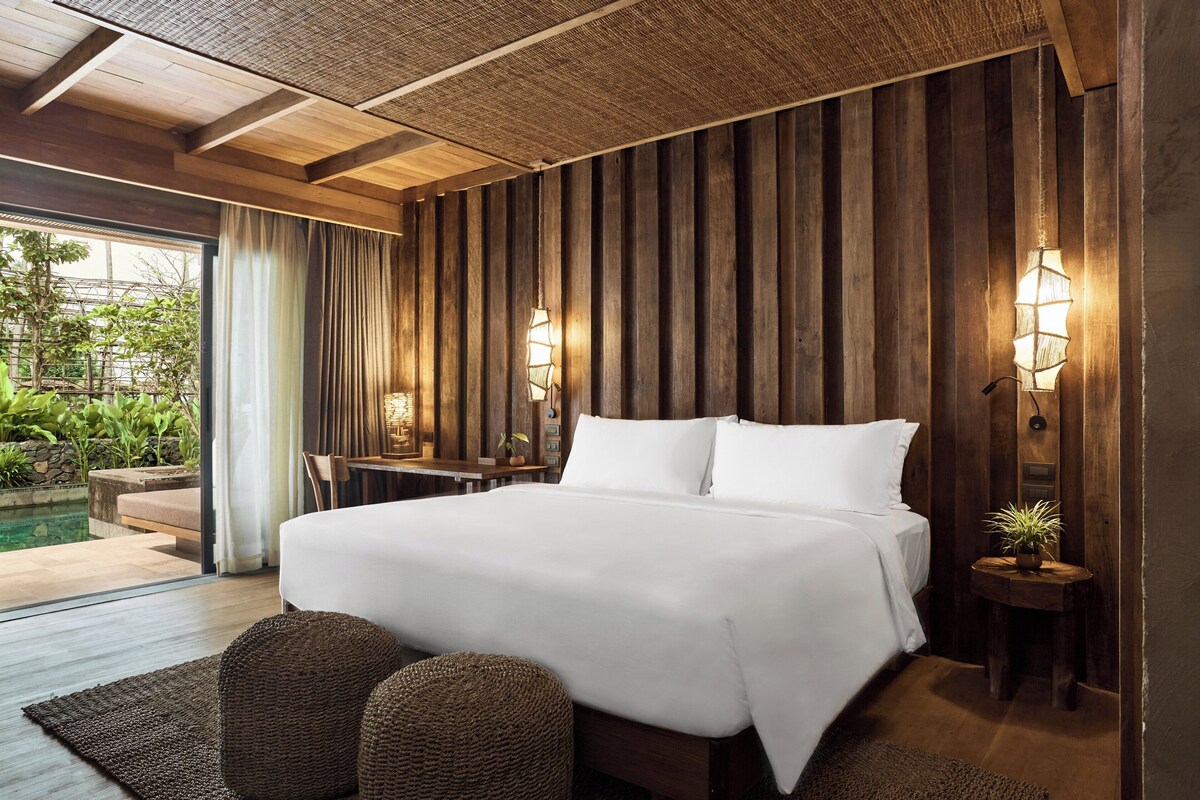 A spacious bedroom with a large bed, wooden panel walls, two bedside lamps, a small table with chairs, and a view of an outdoor pool area.