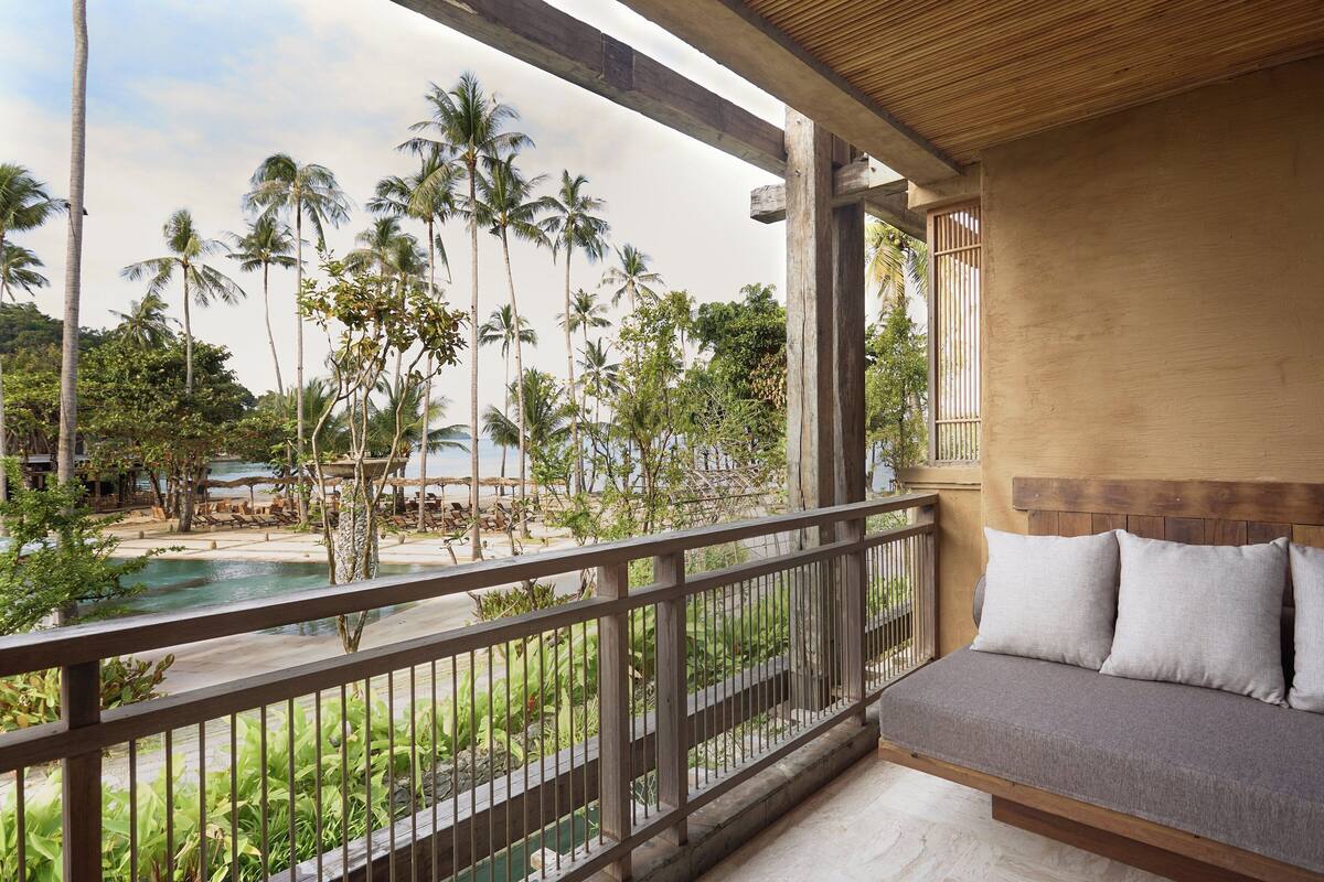 A balcony with a view of a pool and palm trees, a wooden bench, and a wooden ceiling.