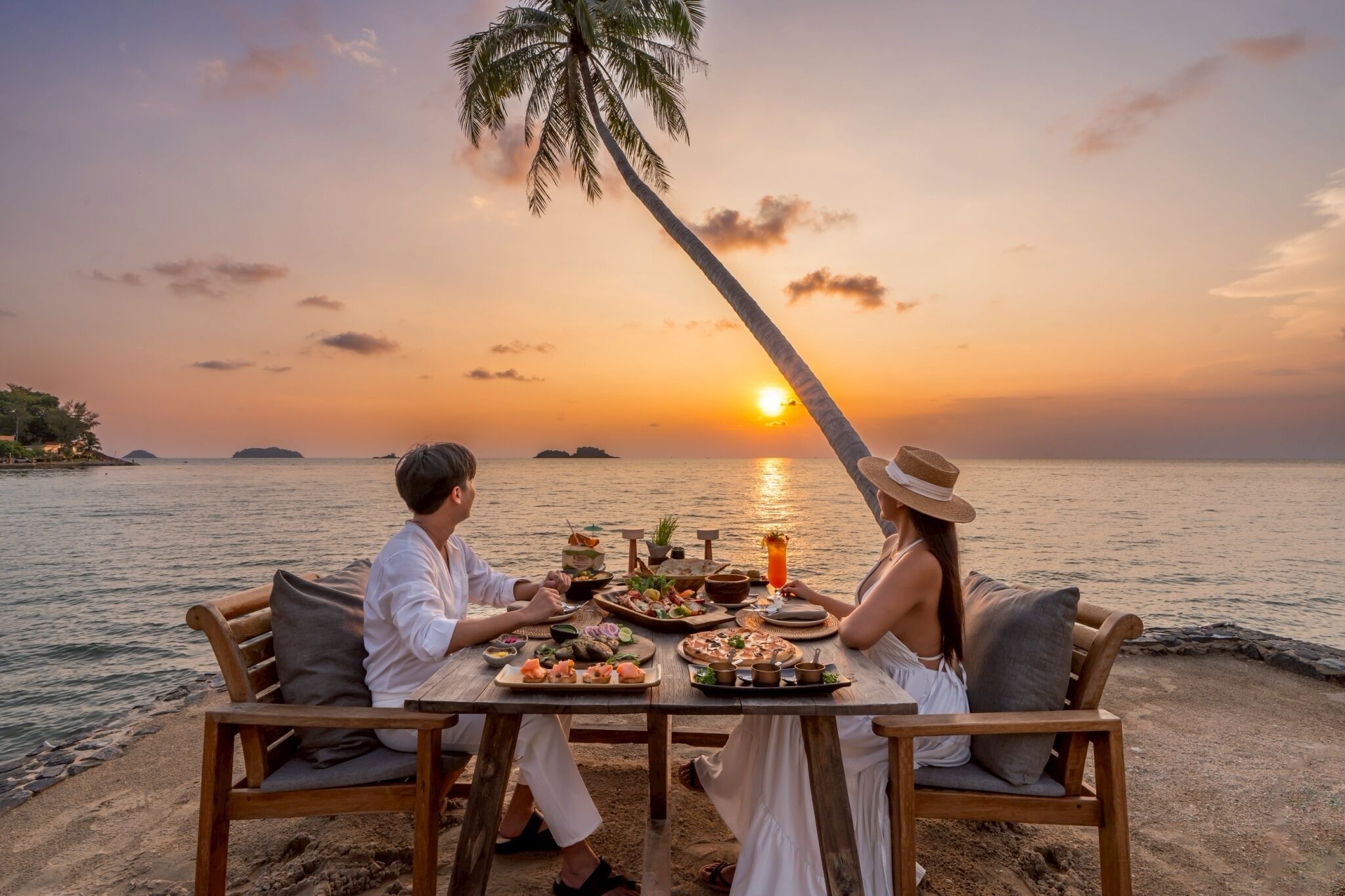 Una pareja cenando en una mesa de madera con vista al atardecer, rodeada de vegetación tropical y una palmera.