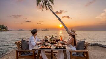 Una pareja cenando en una mesa de madera con vista al atardecer, rodeada de vegetación tropical y una palmera.