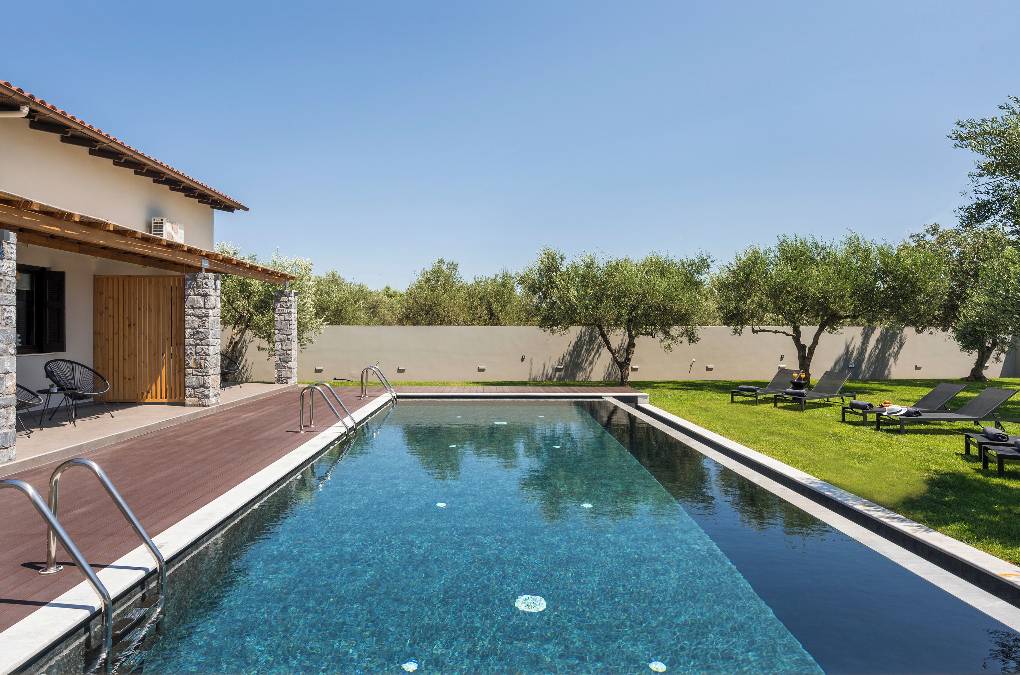 Una piscina con cubierta de madera y sillas de descanso, rodeada por un muro de piedra y árboles.