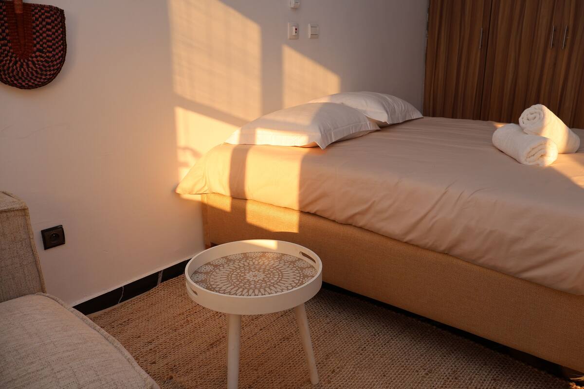 A neatly made bed with white linens and rolled towels, a small round table with a woven top, and a woven rug on the floor.
