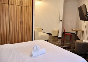 A hotel room with a large bed, a wooden wardrobe, a small white sofa, a red chair, a TV on a stand, and a small table with a remote control. - Casa Hôtel Résidence (Abidjan)
