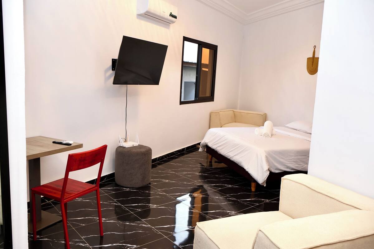 A hotel room with a bed, a TV mounted on the wall, a small table, a red chair, and a grey ottoman.