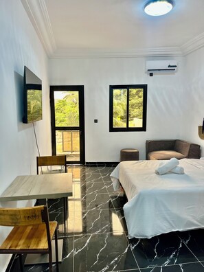 A hotel room with a bed, a desk with a chair, a TV mounted on the wall, and two windows providing a view of greenery. - Casa Hôtel Résidence (Abidjan)