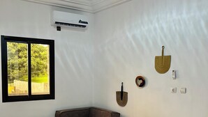 A hotel room with a bed, a brown armchair, a small round ottoman, and a window with a view of greenery. - Casa Hôtel Résidence (Abidjan)