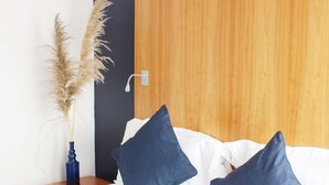 A neatly made bed with white bedding and two dark blue pillows, a wooden headboard, a small wooden nightstand with a blue vase and pampas grass, and a white wall. - Au Bon Coin du Lac (Mimizan)