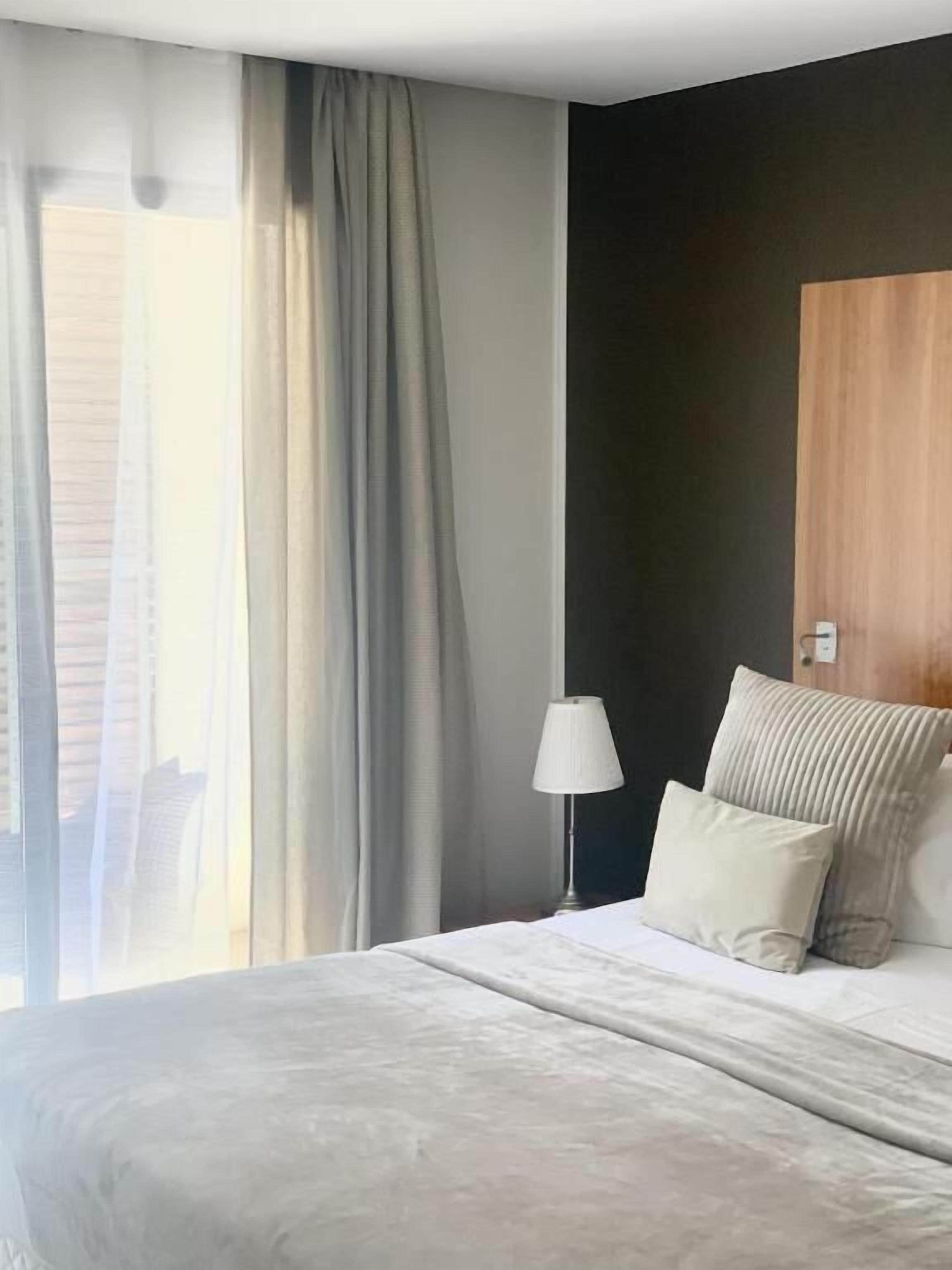Una habitación de hotel moderna con una cama grande, una lámpara de noche y vistas de la ciudad a través de cortinas translúcidas.