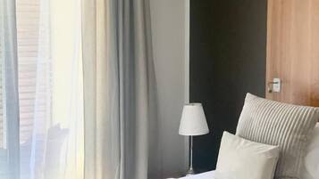 Una habitación de hotel moderna con una cama grande, una lámpara de noche y vistas de la ciudad a través de cortinas translúcidas.