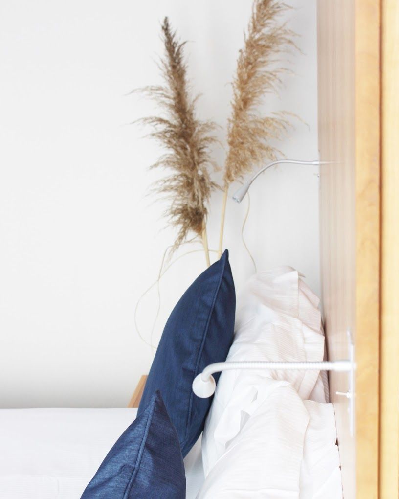 A neatly made bed with white and blue bedding, a white pillow, and two pampas grass plants on a white wall.