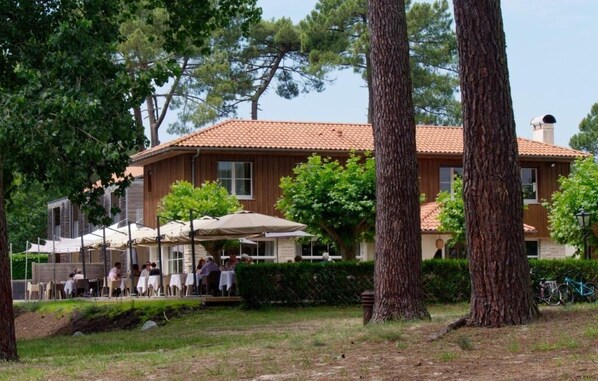 A building with a tiled roof, outdoor seating with umbrellas, and trees in the foreground.