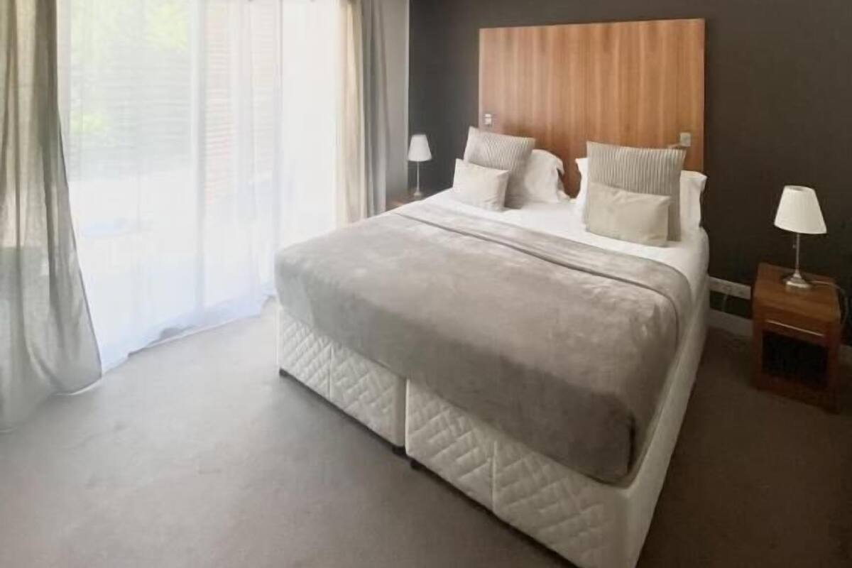 Ett modernt hotellrum med en stor säng, ett träfärgat sänggavel och utsikt över omgivningarna genom tunna, genomskinliga gardiner.