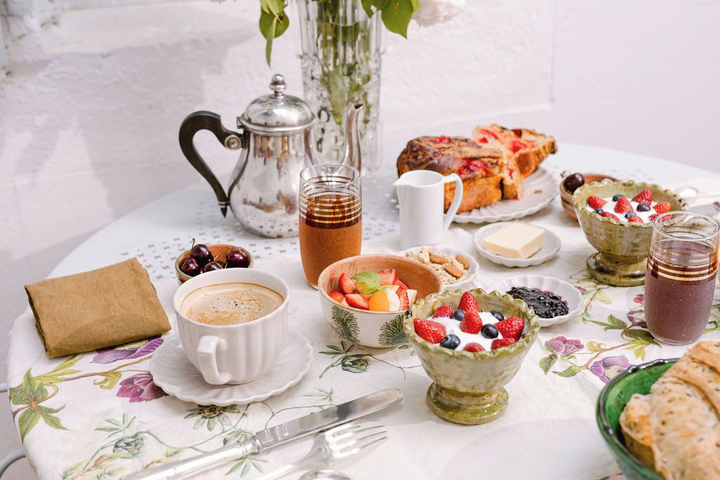 Une table mise pour le petit-déjeuner, avec une théière, une tasse de café, un panier de fruits et des viennoiseries, le tout posé sur une nappe à motifs floraux.