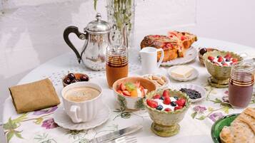 Une table mise pour le petit-déjeuner, avec une théière, une tasse de café, un panier de fruits et des viennoiseries, le tout posé sur une nappe à motifs floraux.