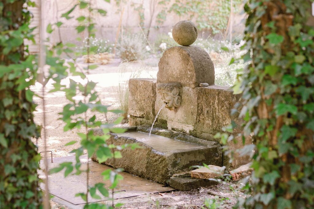 A stone fountain with a lion head spout, surrounded by greenery.