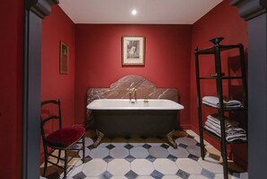 Een badkamer met een vrijstaand bad, een stoel, een plank met handdoeken en een foto in een lijst aan de muur.