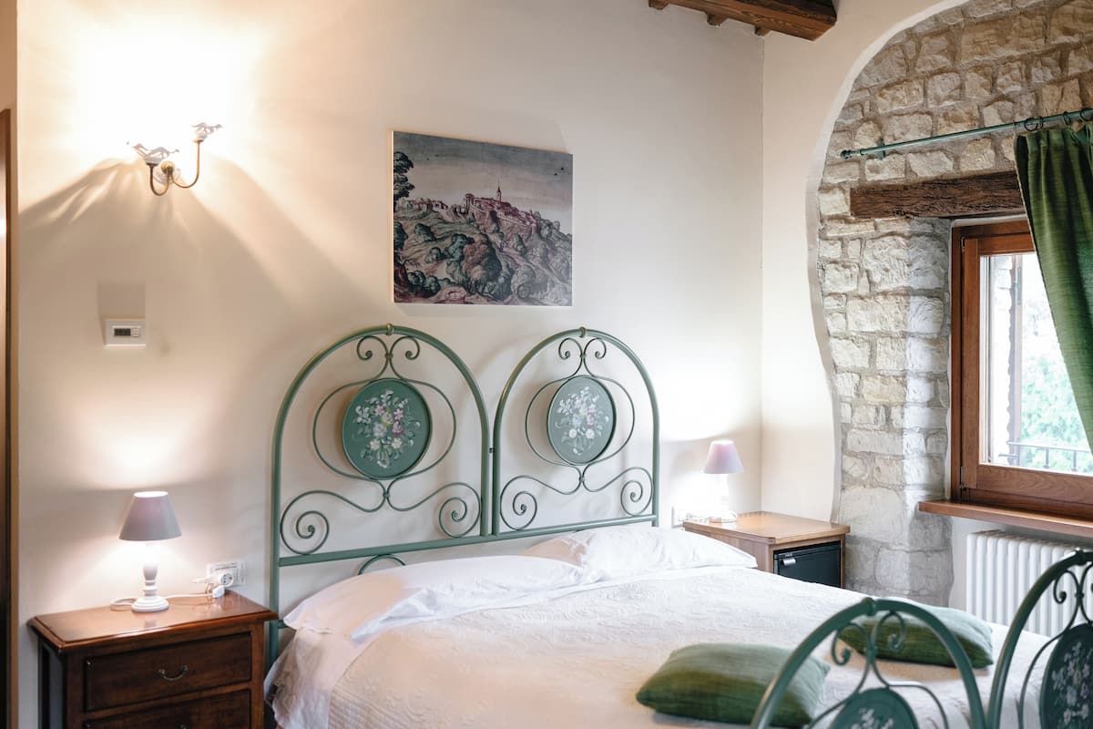 Ein Schlafzimmer mit Steinsandsteinwand, einem Bett mit Metallkopfteil, einem Nachttisch, einer Lampe und einem Fenster mit Vorhängen.