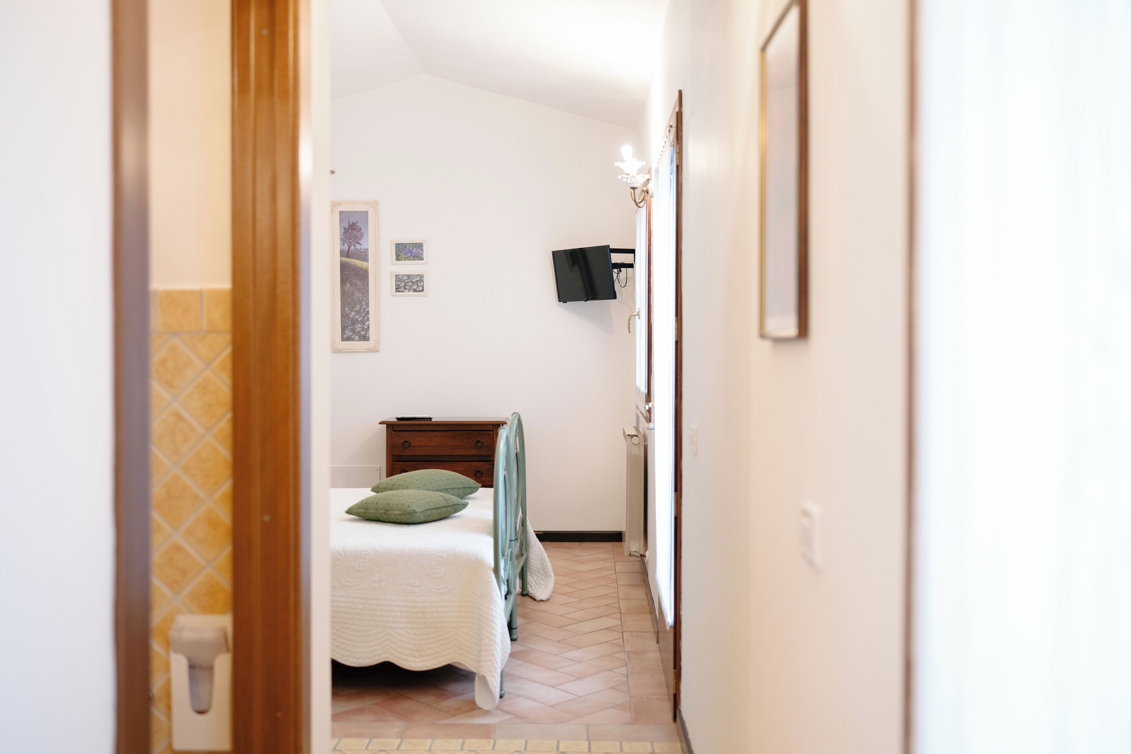 Um corredor estreito que leva a um quarto com uma cama, cômoda e televisão fixada na parede.