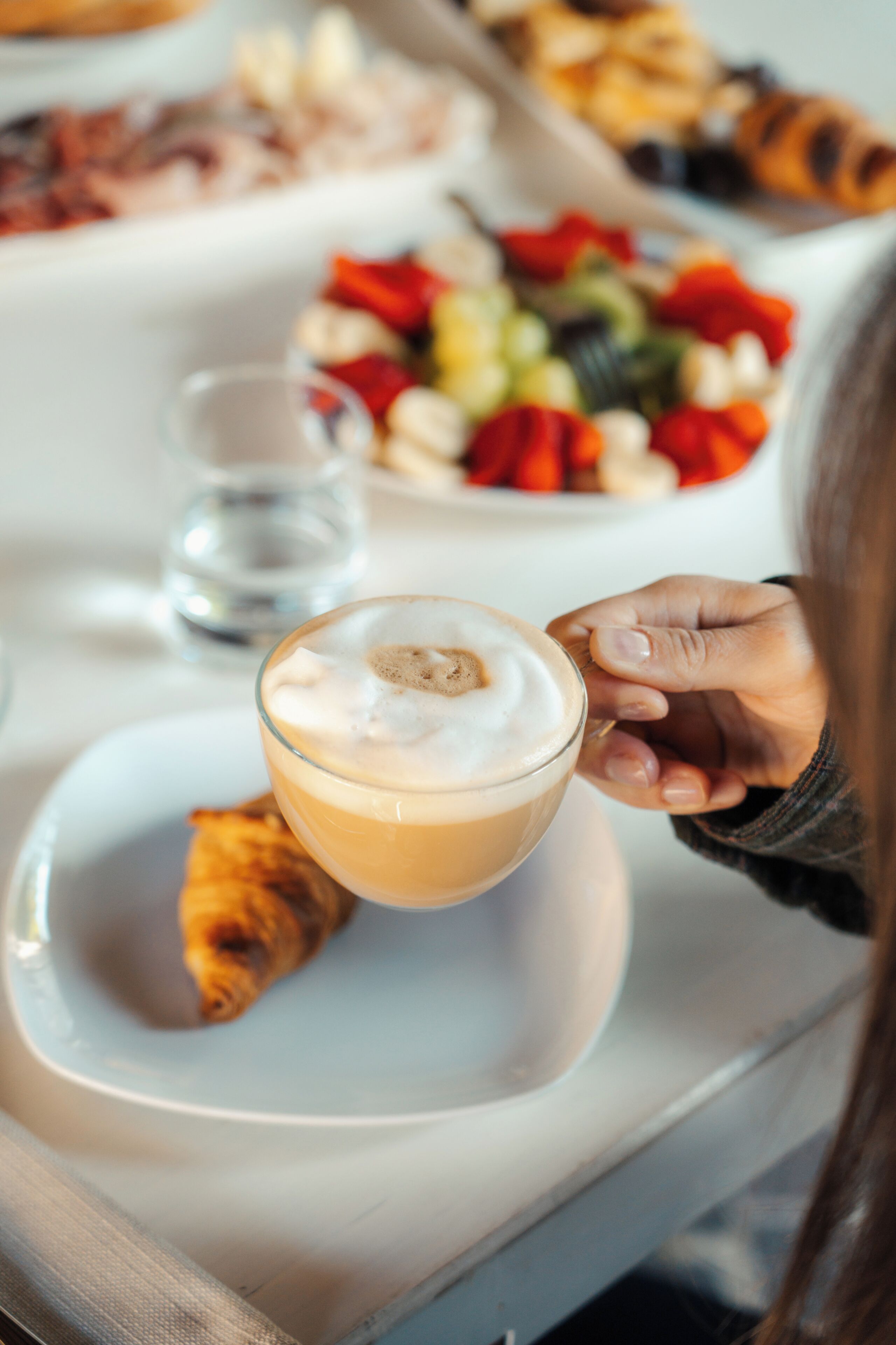 A person holding a cup of coffee with a pastry on a plate, with a table setting in the background.