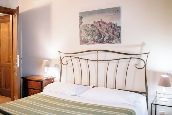 A neatly made bed with a metal headboard, a wooden nightstand with a lamp, and a painting on the wall. - Le Fontanelle Country House (Sant'Ippolito)