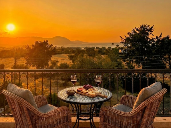 A balcony with wicker chairs, a round table with food and wine, and a sunset view.