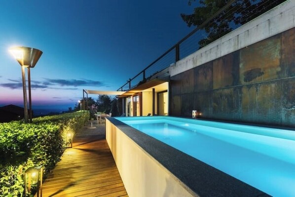 A modern outdoor swimming pool with a wooden deck and ambient lighting, surrounded by greenery and a concrete wall. - House on the Langhe - Luxury private Pool, Sauna and Jacuzzi (Diano D'alba)