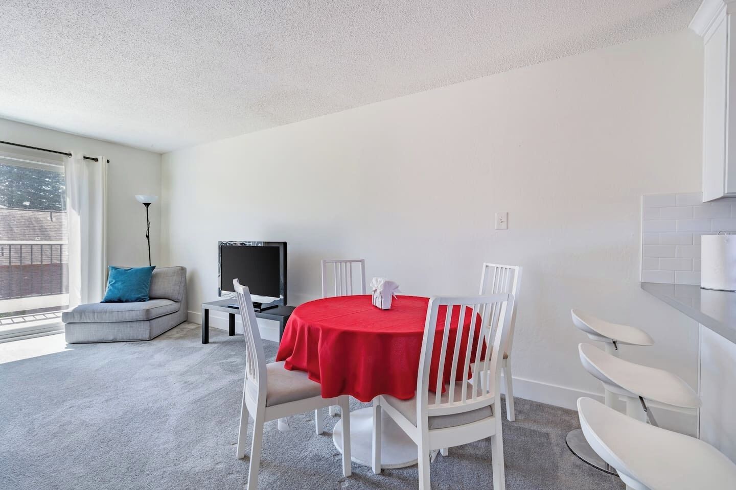 A modern living room with a round table covered in a red tablecloth, white chairs, a grey sofa, a flat-screen TV, and a large window offering a view of the city.