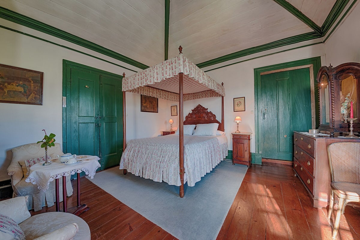 A four-poster bed with a canopy, a wooden dresser, a chair, and a small table with a plant.