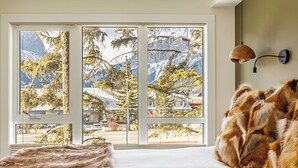 Ein Schlafzimmer mit großem Fenster und Blick auf die Berge, einem Bett mit Felldecke und einer Wandlampe.