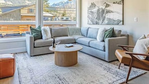 Un salon moderne avec un canapé d’angle, une table basse ronde et une vue sur les montagnes grâce à de grandes fenêtres.