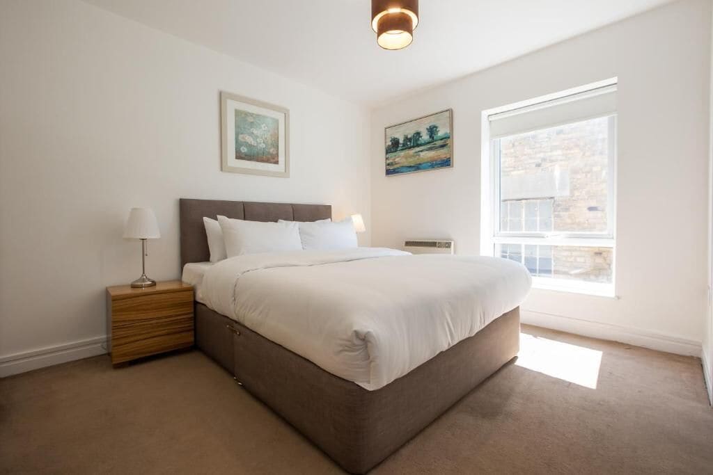 A bedroom with a large bed, a nightstand, a wall-mounted lamp, and two framed pictures.