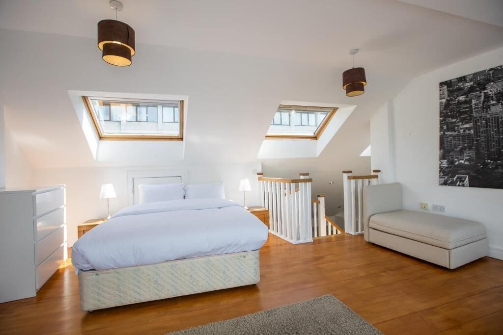 A bedroom with a bed, a nightstand, a dresser, a window, a skylight, a sofa, and a cityscape painting.