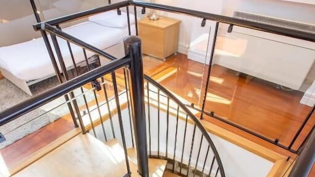 Eine Wendeltreppe mit Glasgeländern und Holzhinweisen führt zu einem Schlafzimmer mit Bett und Nachttisch.