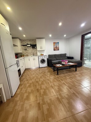 A modern kitchen with white cabinets, a washing machine, and a dining area with a sofa and coffee table.