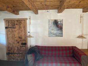 Une pièce avec une porte en bois, un canapé à carreaux rouges et gris, un tableau au mur et deux lampes suspendues.
