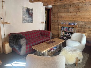 Un salon chaleureux avec un canapé à carreaux rouges, une table basse en bois, un fauteuil blanc, un chien blanc et duveteux, et une bibliothèque remplie de livres.