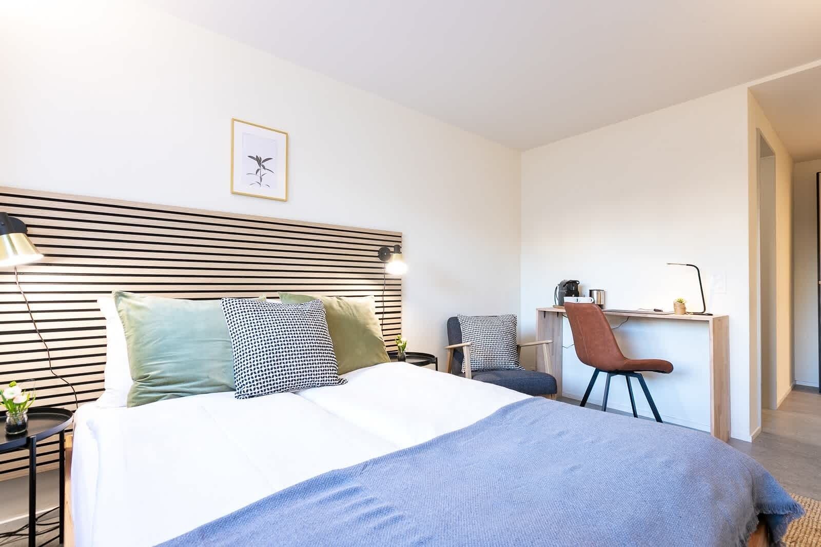 A modern hotel room with a bed, bedside tables, a desk, and a chair.
