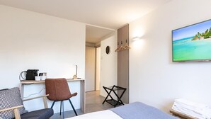 Ein Hotelzimmer mit einem Bett, einem Schreibtisch mit Stuhl, einem Fernseher der eine Strandansicht zeigt und einem an der Wand befestigten Handtuchhalter.