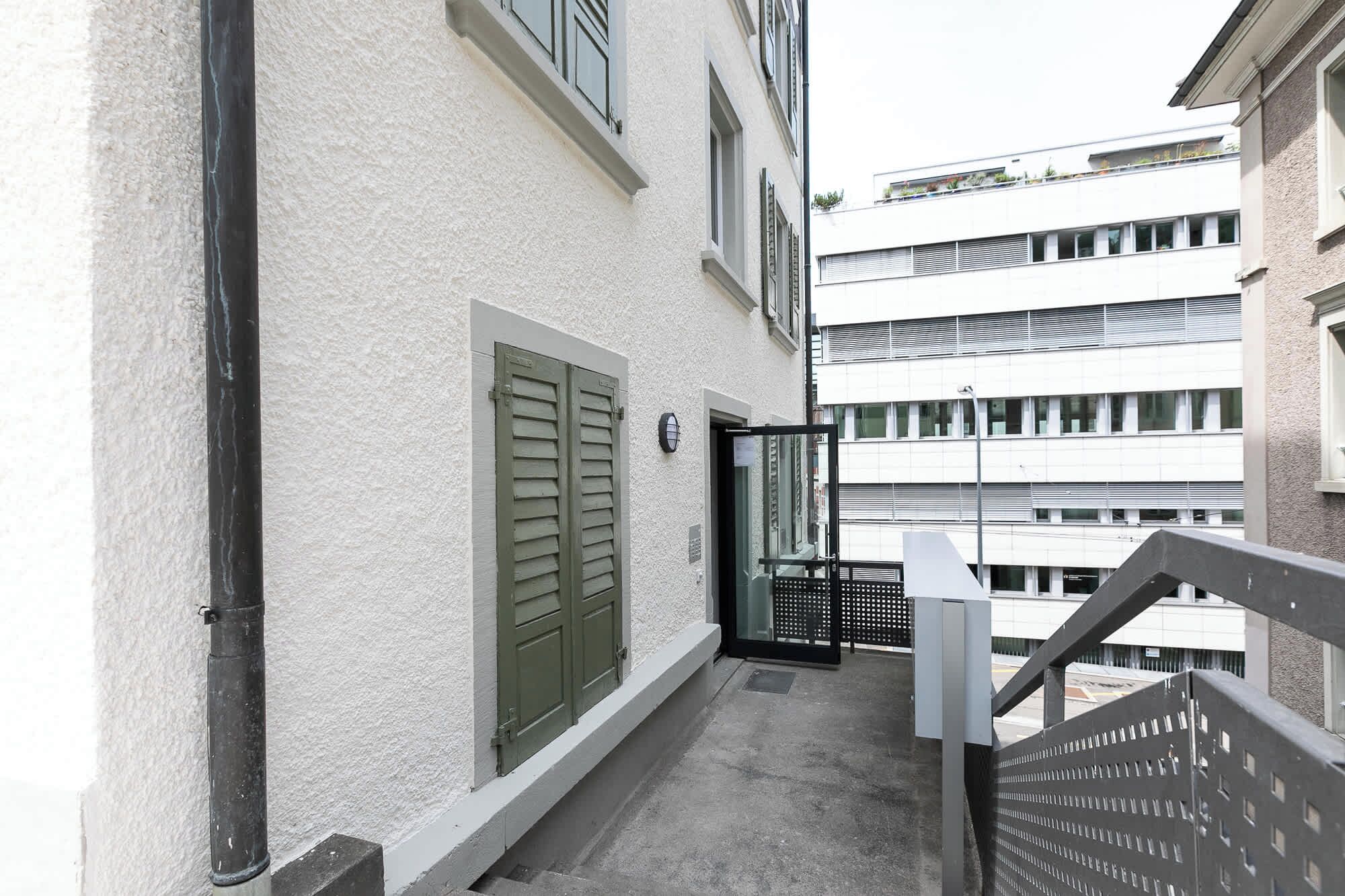 En smal balkong med ett metallstaket, en vit byggnad med fönsterluckor och en hög byggnad i bakgrunden.