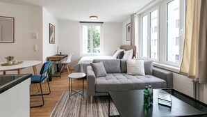 Una habitación moderna y bien iluminada con un sofá gris, una pequeña mesa redonda, un escritorio con una silla y una cama con cabecera.