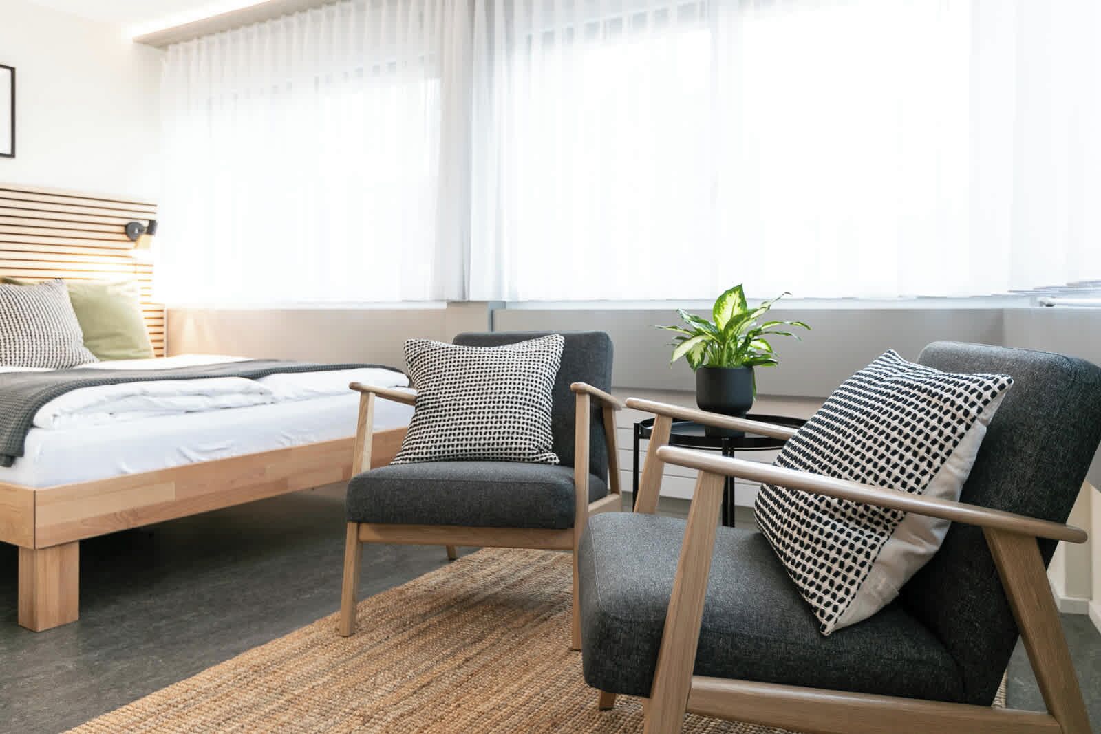 Ein Schlafzimmer mit einem Bett, zwei Sesseln, einem kleinen Tisch mit einer Pflanze und einem Fenster mit Vorhängen.
